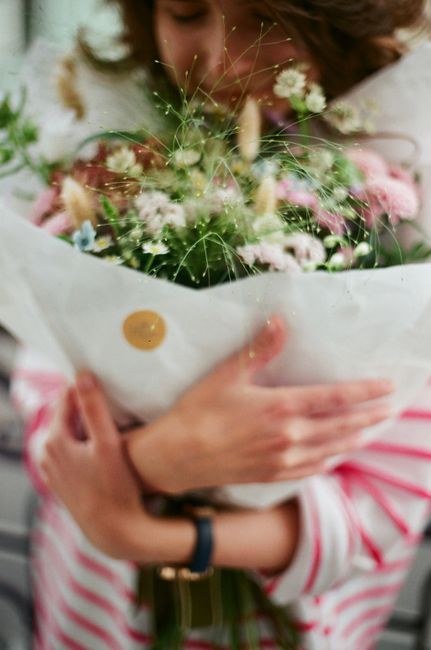A person holding a vibrant bouquet arrangement of fresh flowers, featuring pink roses, white baby's breath, and green foliage, wrapped in white paper with a small round sticker. The arrangement includ