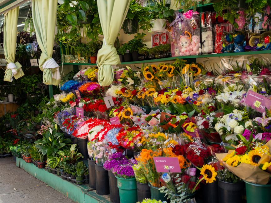 A vibrant display of fresh floral arrangements outside a flower shop, featuring a variety of bouquets with bright sunflowers, roses, and lilies wrapped in clear cellophane and decorative paper. The bo