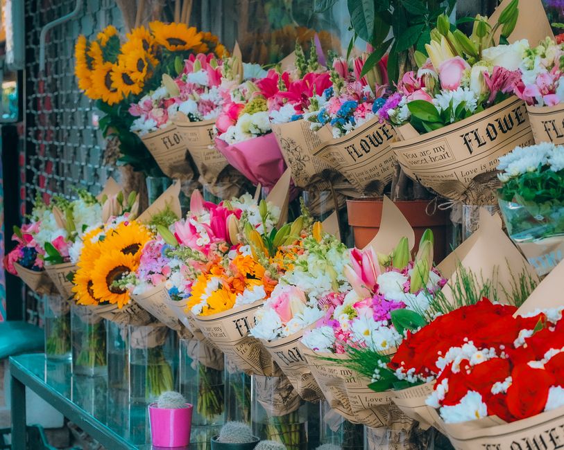 A vibrant display of fresh floral bouquets arranged on a glass shelf, featuring a variety of flowers including yellow sunflowers, pink and white lilies, pink roses, blue and purple blooms, and red car