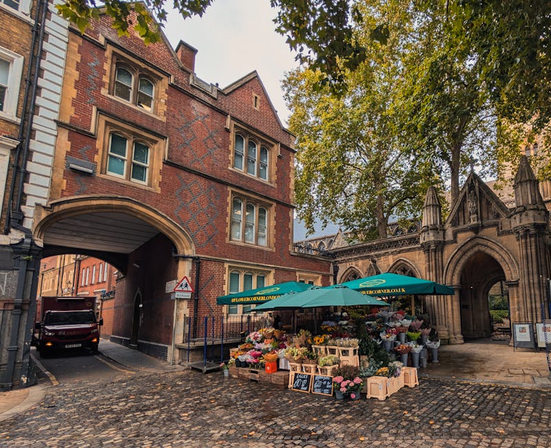 A bustling flower stall situated on a cobblestone street next to an old brick building with an arched passageway, displaying a wide selection of fresh, vibrant flowers including roses, peonies, and li