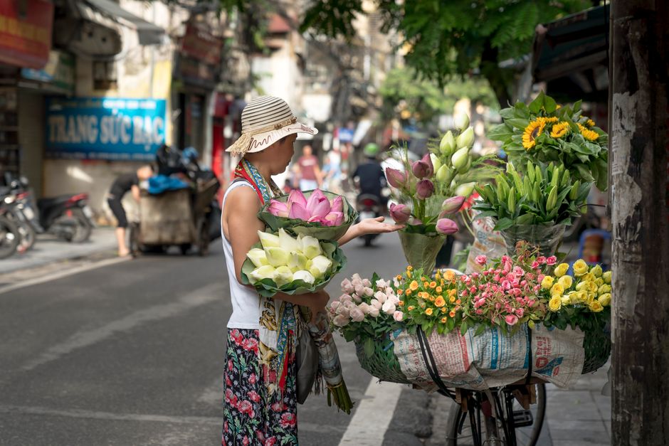 A woman wearing a wide-brimmed straw hat, a sleeveless white top, and a floral skirt is standing on a busy urban street, selecting flowers from a floral vendor's cart. She holds a bouquet of white and