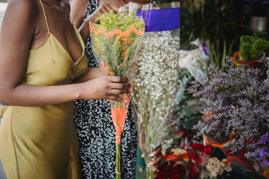 A woman in a yellow satin dress holds a bouquet of mixed flowers wrapped in clear cellophane with orange and pink accents, featuring green hydrangeas and purple statice, in a flower shop. The bouquet